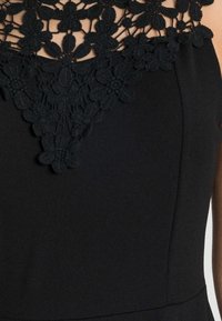 Svart klänning med en blommig spetsdesign vid halsen och en mjuk, slät textur på bysten.