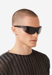 Čierne slnečné okuliare s obtočujúcim dizajnom, tmavými sklami a štýlovým rámom. Nosené osobou s krátkym strihom vlasov a pruhovanou čiernou košeľou.