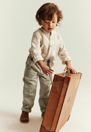 Peuter in beige overhemd en broek staat en houdt een grote bruine vintage koffer vast tegen een lichte achtergrond.