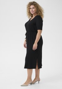 Black midi dress with short sleeves, a V-neck, tie waist detail, and side slits. Made from a textured fabric with a smooth finish.