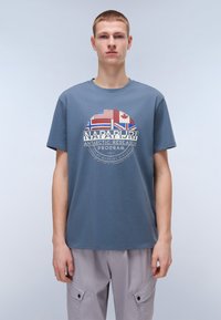 T-shirt en coton bleu avec un graphisme circulaire comprenant des drapeaux, du texte et un logo. Design à col rond et manches courtes. Coupe simple et décontractée.