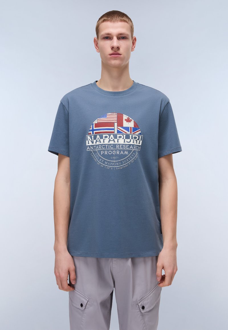 T-shirt en coton bleu avec un graphisme circulaire comprenant des drapeaux, du texte et un logo. Design à col rond et manches courtes. Coupe simple et décontractée.