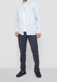 Muž v světle modré košili s vzorem, bílem tričku s potiskem, tmavě modrých károvaných kalhotách a námořnických semišových mokasínách na bílém pozadí.