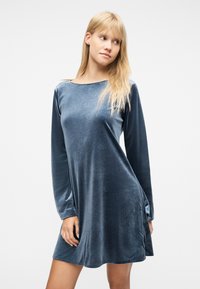 Νεαρή γυναίκα με μακριά ξανθά μαλλιά, φοράει ένα φαρδύ, μακρυμάνικο μπλε βελούδινο φόρεμα, κοιτάζει προς τα δεξιά της πάνω σε απλό φόντο.