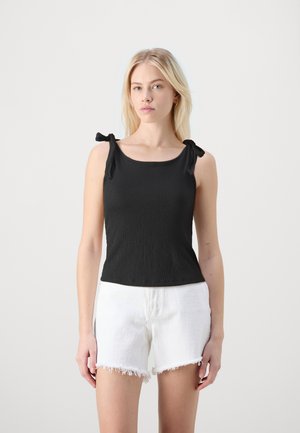 Tops noir sans manches en tissu texturé, avec des bretelles nouées aux épaules et un décolleté arrondi, associé à un short en denim blanc effiloché.
