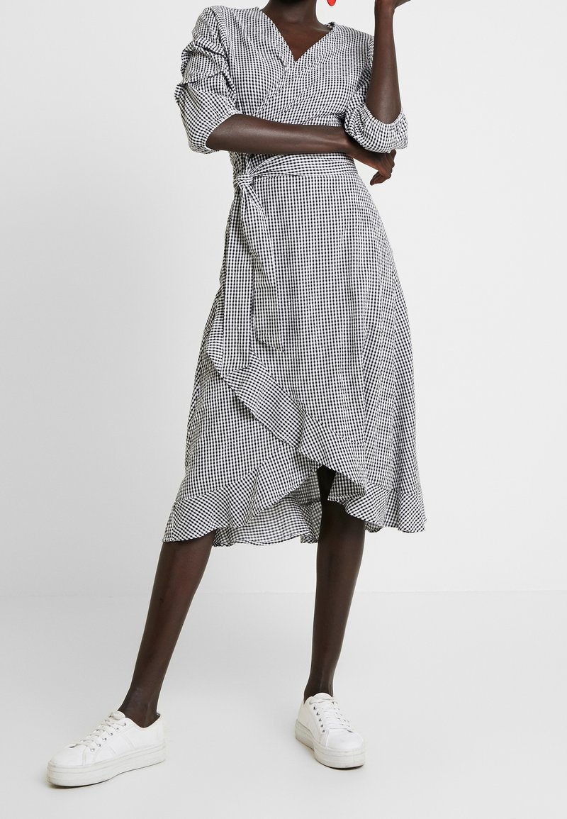 Robe portefeuille à carreaux vichy noir et blanc avec manches bouffantes, ourlet asymétrique et accents à volants, portée avec des baskets blanches.