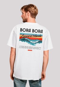 Weißes Baumwoll-T-Shirt mit einem grafischen Druck auf der Rückseite, der "Bora Bora" und farbenfrohe Wellenmuster in Blau, Orange und Türkis zeigt.
