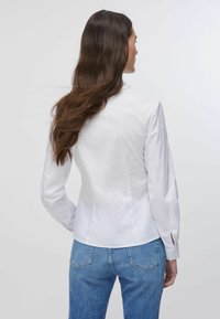 Chemise blanche à manches longues avec une coupe ajustée, présentant un léger brillant et des accents découpés aux poignets. Associée à un jean en denim bleu.