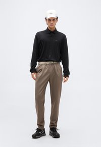 Άνδρας που φοράει λευκό καπέλο, μαύρο μακρυμάνικο πουκάμισο, μπεζ παντελόνι με ζώνη και μαύρα αθλητικά παπούτσια, στέκεται μπροστά σε απλό φωτεινό φόντο.