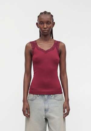 Femme aux cheveux tressés portant un haut bordeaux sans manches avec bordure en dentelle et un jean ample bleu clair sur un fond uni.