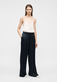 Νεαρή γυναίκα όρθια με το ένα χέρι στην τσέπη, φορώντας λευκή αμάνικη μπλούζα με δαντέλα και φαρδιά μαύρη παντελόνια με λευκά παπούτσια.