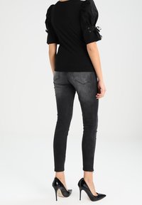Donna che indossa un top nero con maniche a sbuffo, jeans skinny neri sbiaditi e scarpe con tacco nere, in piedi con la schiena rivolta verso la fotocamera.