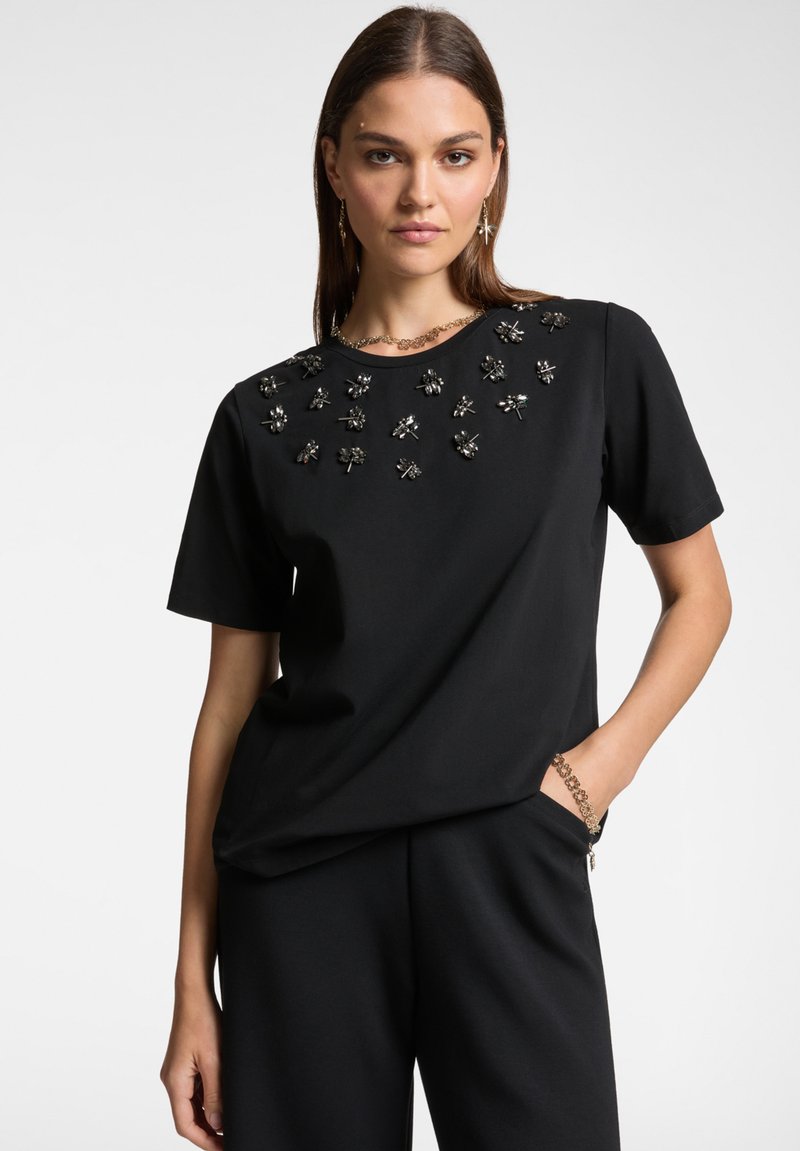 Top nero a maniche corte con scollatura rotonda. Presenta decorazioni sul petto che richiamano disegni floreali. Tessuto morbido e vestibilità comoda.