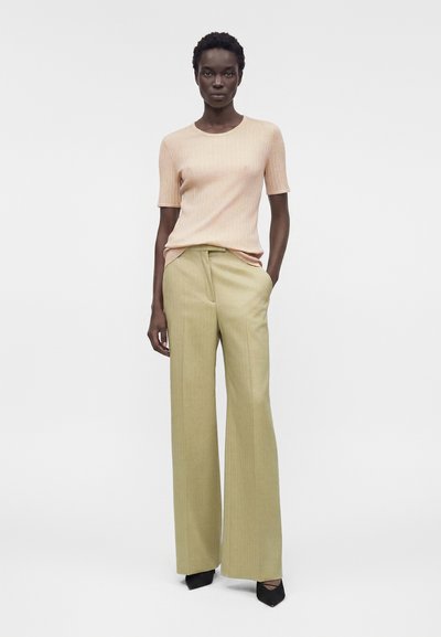 Γυναίκα που φοράει ροδακινί ριμπ πλεκτή κοντομάνικη μπλούζα, μπεζ ψηλόμεσο παντελόνι με φαρδιά μπατζάκια και μαύρα παπούτσια με μυτερή μύτη, στέκεται με το ένα χέρι στην τσέπη.