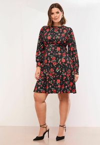 Juodas gėlių suknelė ilgomis rankovėmis, puošta raudonomis gėlėmis ir žaliais lapais, širdaus sijono ir elastinga juosmena. Juodų aukštakulnių batų pora.