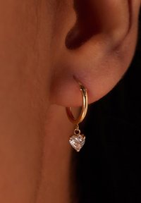 Gouden hoepeloorbel met een hartvormige heldere edelsteen die hangt. Voorzien van een gepolijste afwerking en een delicaat scharnier sluitmechanisme.