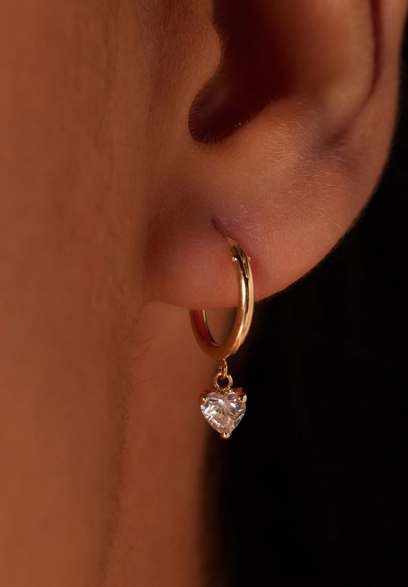 Gouden hoepeloorbel met een hartvormige heldere edelsteen die hangt. Voorzien van een gepolijste afwerking en een delicaat scharnier sluitmechanisme.