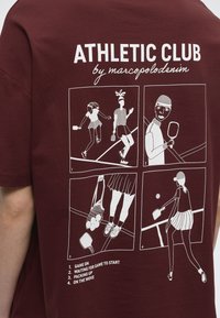 Σχέδια με λευκές γραμμές σε τέσσερα πάνελ σε μπλούζα μπορντό δείχνουν άτομα που παίζουν και μαζεύουν εξοπλισμό για ένα άθλημα με ρακέτες, υπό τον τίτλο "ATHLETIC CLUB by marcopolodanim."