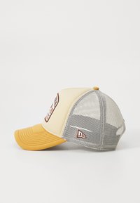 Gorra trucker amarilla y beige con parte trasera de malla, visera curva y parche de logo bordado en la parte frontal. Cierre ajustable en la parte trasera.