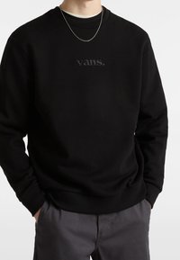 Felpa nera in tessuto morbido con scollo rotondo e logo "vans." ricamato sul petto. Abbinata a pantaloni grigi.