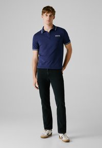 Marineblauwe poloshirt met witte accenten, een enkel borstlogo en korte mouwen, gedragen met donkere jeans en lichtgekleurde sneakers.