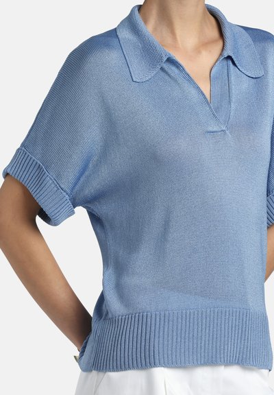 Pulover polo tricotat cu mâneci scurte, de culoare albastru deschis, cu manșete și tiv striate, purtat nes tucked peste pantaloni albi.