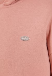 Un hoodie rose avec une texture douce présente un petit patch en argent ovale portant l'inscription "Simulator". Le design comprend une coupe décontractée et une capuche.