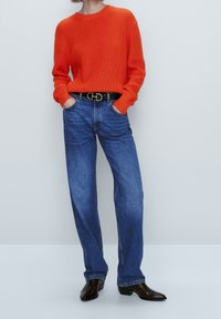 Pull en tricot côtelé orange avec encolure ronde, associé à un jean bleu droit et une ceinture noire avec accessoires en métal argenté.