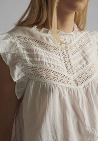 Femme portant une blouse blanche avec des détails en dentelle et des manches volantées, vue de la nuque à la taille sur fond uni.