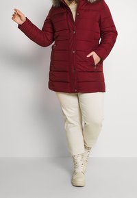 Bordeaux pufferjacka med pälsfodrad huva, med dragkedjestängning, sidofickor och ett kviltat mönster; kombinerad med krämfärgade byxor och snörskor.