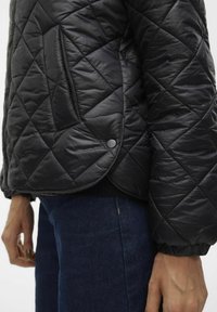 Giacca nera trapuntata con finitura lucida, motivo a rombi, tasca laterale e polsini elasticizzati. Abbinata a jeans di denim scuro.