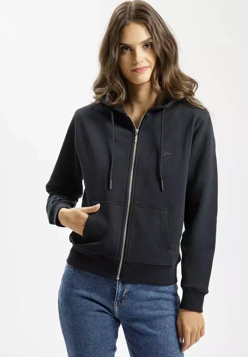Czarna bluza z kapturem na zamek, wykonana z miękkiego materiału, z kieszenią z przodu, regulowanymi sznurkami oraz ściągaczami przy rękawach. Noszona z niebieskimi dżinsami.