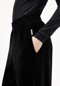 Schwarze Samthose mit einem elastischen Bund, seitlichen Taschen und einem kleinen Etikett an der Tasche. Glatte Textur und lockerer Schnitt.