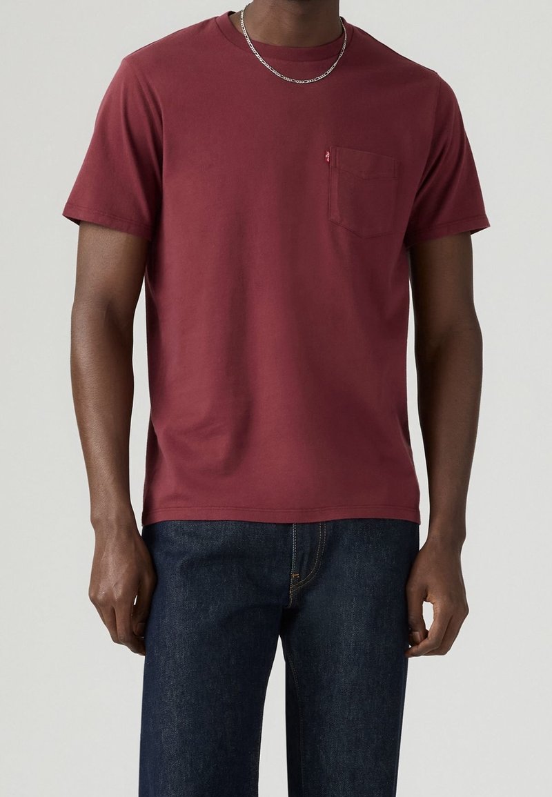 T-shirt bordeaux à manches courtes en coton, avec une poche poitrine et un col rond. Porté avec un jean en denim bleu.