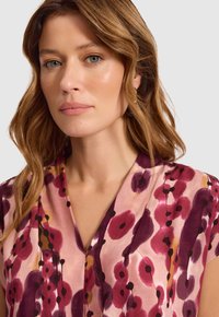 Kobieta z falującymi, jasnobrązowymi włosami, nosząca kolczyki w kształcie koła oraz bluzkę w różowo-fiołkowy kwiatowy wzór, na tle jasnego, jednolitego tła.