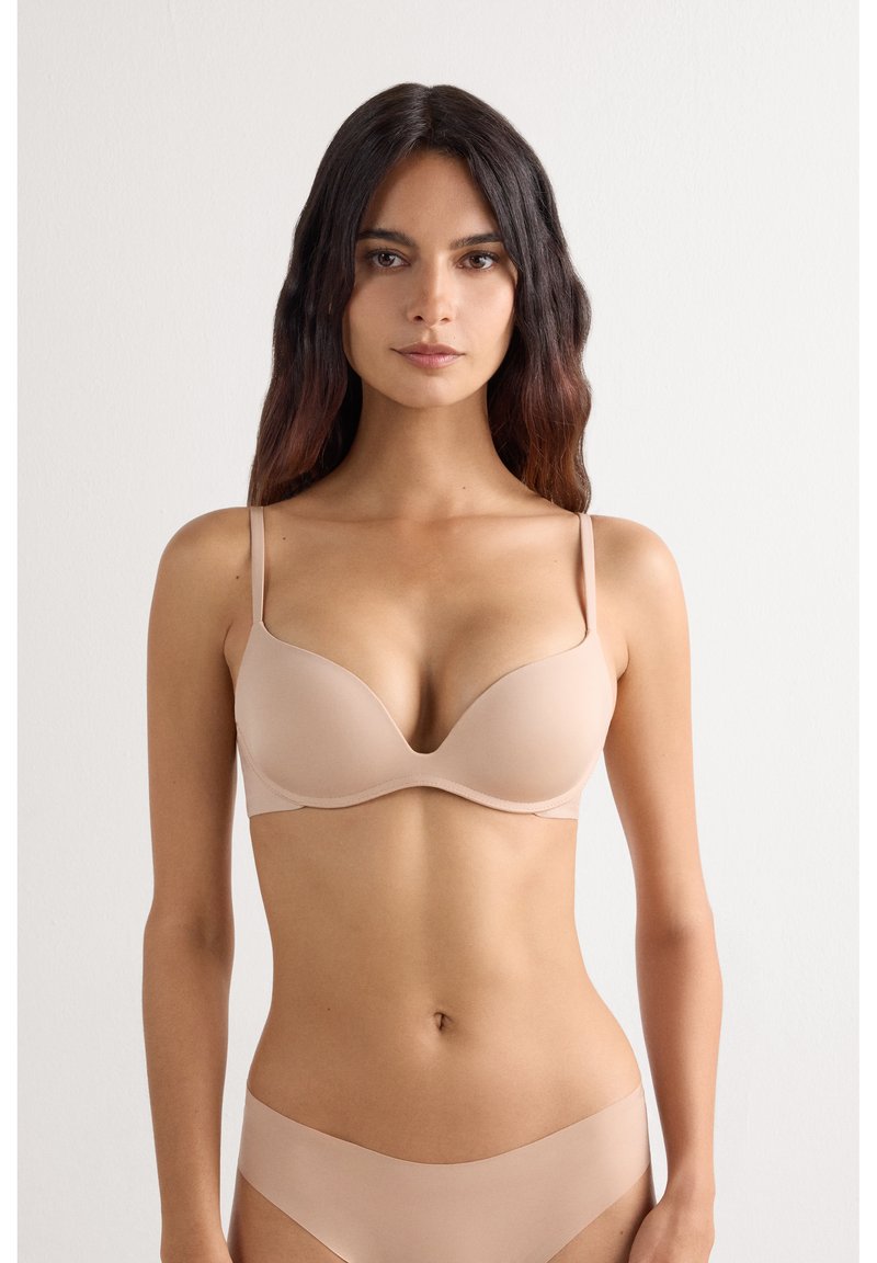 Femme aux longs cheveux foncés portant un soutien-gorge couleur chair et une lingerie assortie sans coutures sur un fond blanc uni.
