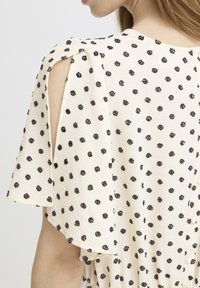 Femme portant un chemisier crème à pois noirs, avec des manches fendues ouvertes et des boutons dans le dos.