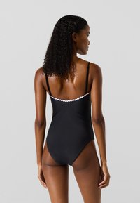 Sieviete ar cirtainiem mitriem matiem, tērpusies melnā viengabala peldkostīmā ar plānām siksniņām un baltu apdari, redzama no aizmugures uz vienkārša fona.