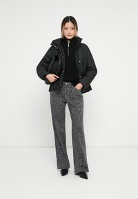 Svart pufferjakke, svart strikkegenser, grå leopardmønstrede bukser, svarte spisse sko, holder en svart veske, står mot en hvit bakgrunn.