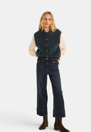 Femme blonde portant un gilet boutonné bleu foncé par-dessus une chemise claire à manches longues, un jean large foncé avec des revers, et des bottines marron.