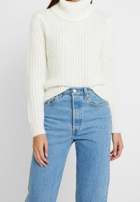 White textured knit turtleneck sweater with ribbed detailing, paired with high-waisted light blue denim jeans. Sweater features long sleeves.