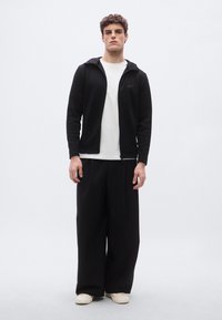 Chaqueta negra con cremallera sobre una camisa blanca, combinada con pantalones negros holgados y zapatos crema. Tejido suave, corte relajado, detalle de logo minimalista.