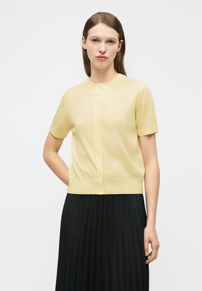 Γυναίκα με ίσια καστανά μαλλιά, ντυμένη με κίτρινη κοντομάνικη ζακέτα με κουμπιά και μαύρη πλισέ φούστα, στέκεται μπροστά σε λευκό φόντο.