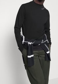 Man med svart långärmad skjorta med liten vit logga nära kragen, mörkgröna byxor och en marinblå och vit tröja knuten runt midjan.