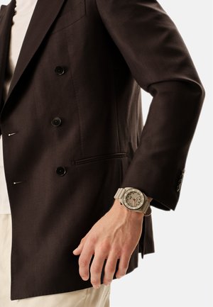 Bras d'un homme portant un blazer marron foncé et une montre-bracelet beige avec un cadran rond et un bracelet en caoutchouc sur un fond clair.