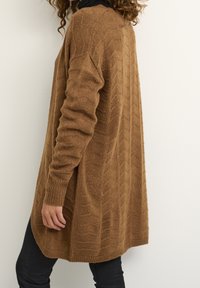 Brauner strukturierter Cardigan mit geriffeltem Muster, langen Ärmeln und lockerer Passform, kombiniert mit schwarzen Hosen. Weiches Strickmaterial.