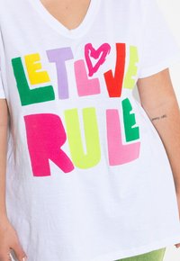 T-shirt blanc à col en V avec un texte coloré et audacieux : "LAISSEZ L'AMOUR RÉGNER" en lettres moelleuses vertes, roses, jaunes, violettes et rouges. Texture douce.