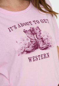 Light pink cotton t-shirt featuring a graphic of brown cowboy boots with floral accents and the text "IT'S ABOUT TO GET WESTERN" in dark maroon.