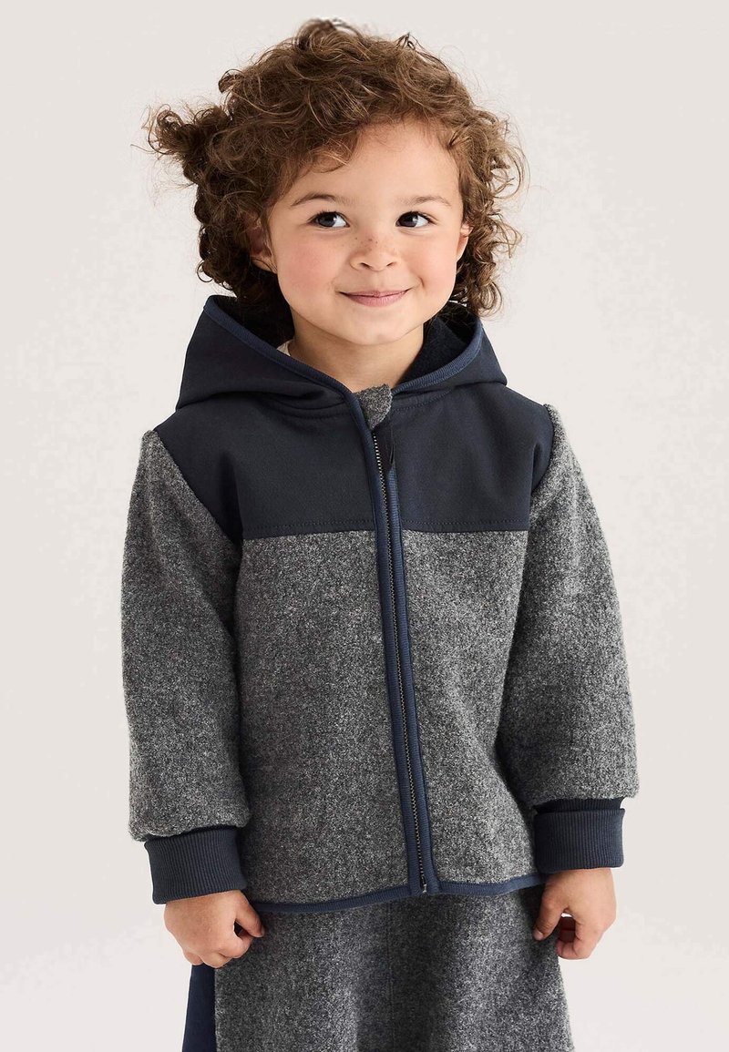 Kleines Kind mit lockigem Haar, das einen grauen und marineblauen Zip-Hoodie trägt, lächelt und schaut leicht zur Seite vor einem schlichten Hintergrund.