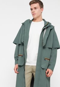 Νεαρός άνδρας φοριέται ένα απαλό πράσινο μακρύ αδιάβροχο με φερμουάρ, μπεζ παντελόνι και λευκή μπλούζα, κοιτάζοντας ελαφρώς πλάγια.